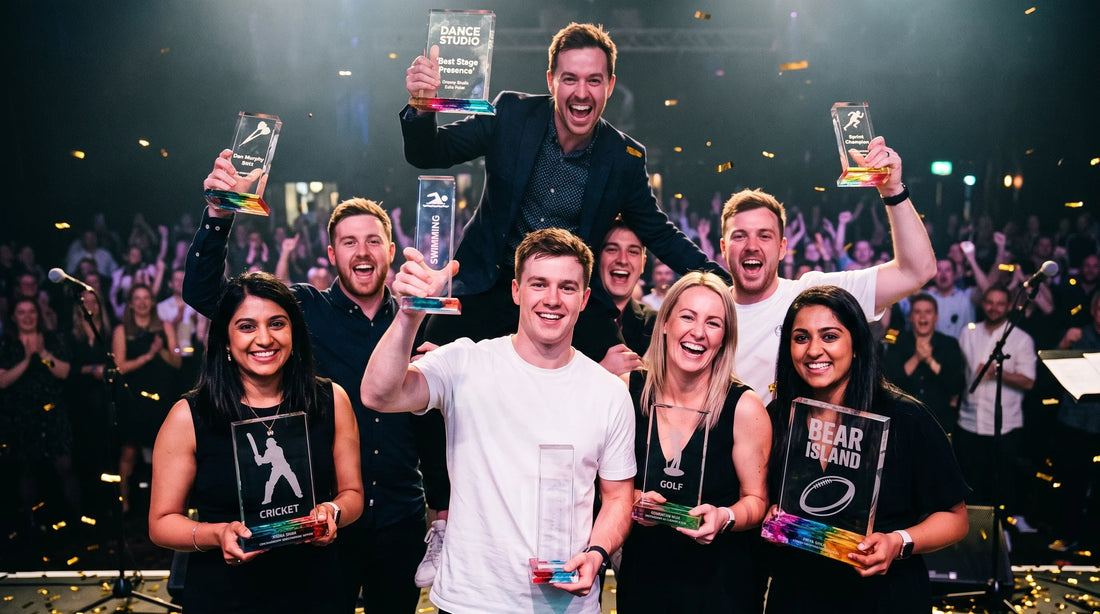 Awards gala night stage celebration, featuring a group of smiling winners holding custom crystal and color-accented trophies—including 'Cricket', 'Golf', 'Dance Studio Best Stage Presence', and 'Swimming'—amidst falling confetti and an applauding audience