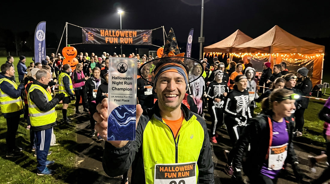 Runner in a witch hat holding a Halloween Night Run Champion acrylic trophy at a nighttime fun run, with costumed runners and a Halloween Fun Run banner behind him