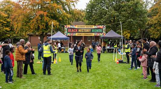 Participants running across the finish line under a Black History Month sport and community banner with cheering spectators on a green park field.