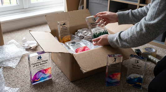 Person unpacking and organizing crystal and acrylic award trophies from a cardboard box on a carpeted floor.